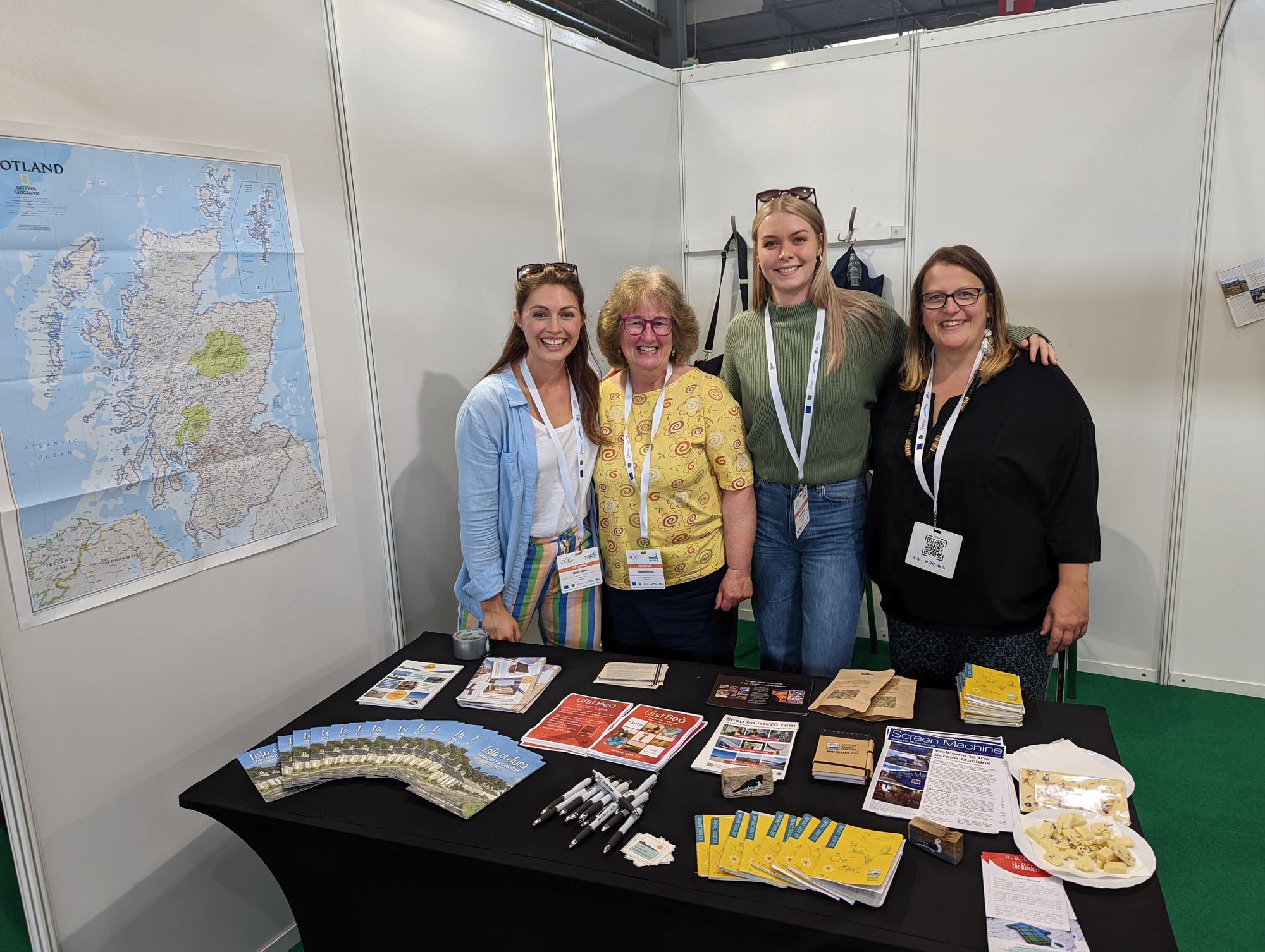 Four people stand smiling behind a table with brochures and pens. Theres a map of Scotland on the wall. They are at an exhibition booth with white walls and a green floor.