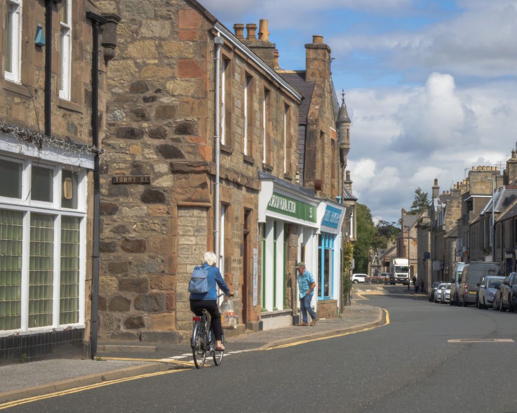 A person wearing a blue jacket cycles down a quiet street lined with stone buildings and shops, while another person walks on the sidewalk under a partly cloudy sky.