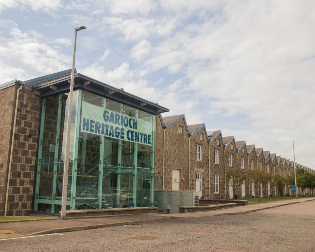 The image shows the exterior of the Garioch Heritage Centre, a modern glass entrance attached to a long, stone building with pitched roofs, under a partly cloudy sky.