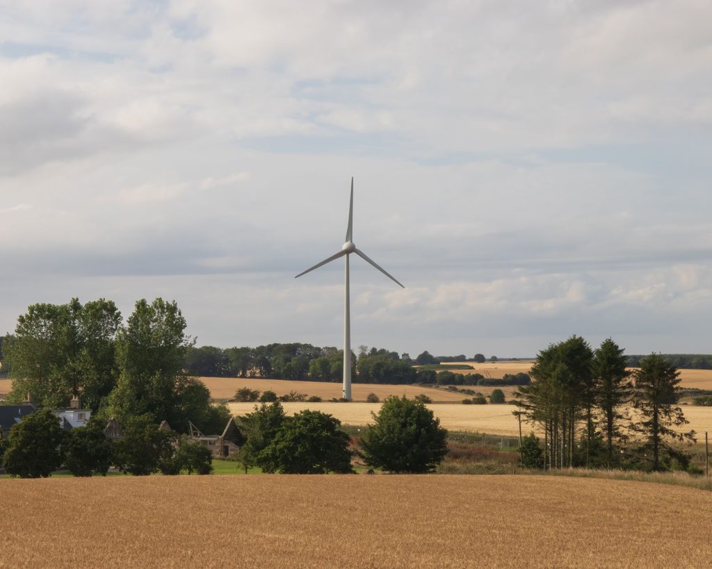 A large wind turbine stands in a golden field surrounded by trees and farmland under a partly cloudy sky. Some houses are partially visible among the trees in the foreground.