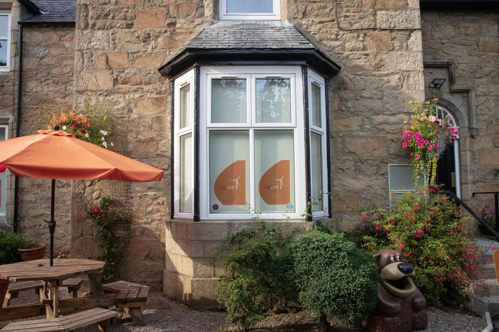 A stone building with a large bay window, orange decals on the glass, hanging flower baskets, a wooden picnic table with an orange umbrella, and a bear-shaped planter in the garden.