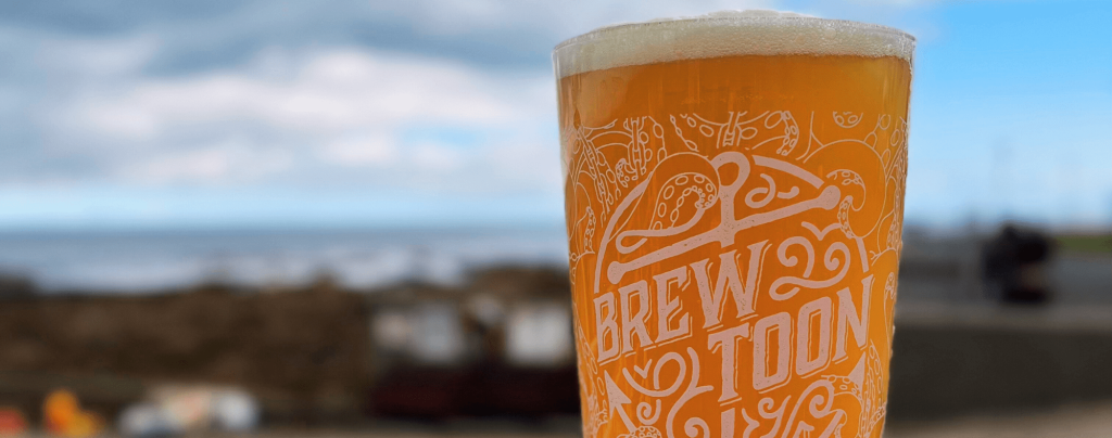 A pint glass of beer with Brew Toon printed on it is held up outdoors, with a blurred background of the coastline, cloudy sky, and sea.