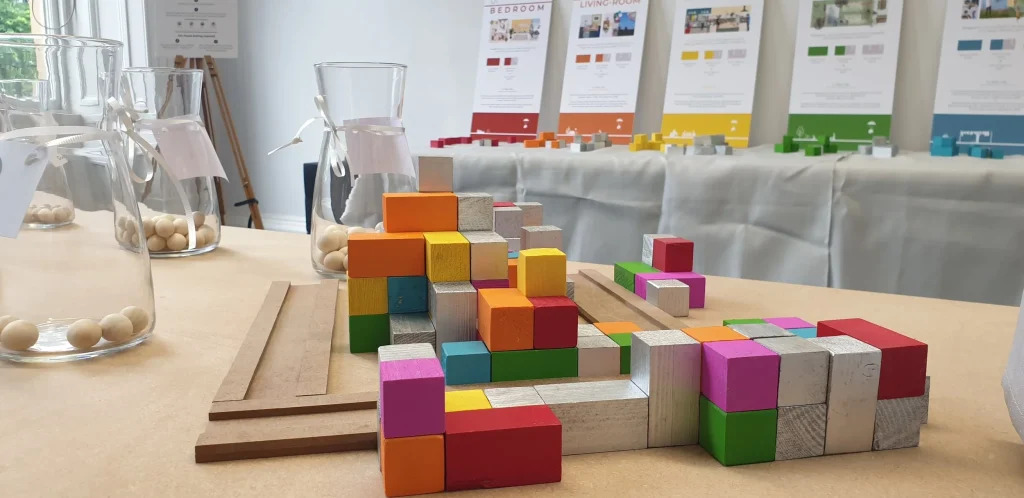 Colorful wooden blocks are arranged on a table, with more blocks scattered nearby. In the background, posters with diagrams and jars filled with wooden beads are visible, suggesting a workshop or educational setting.
