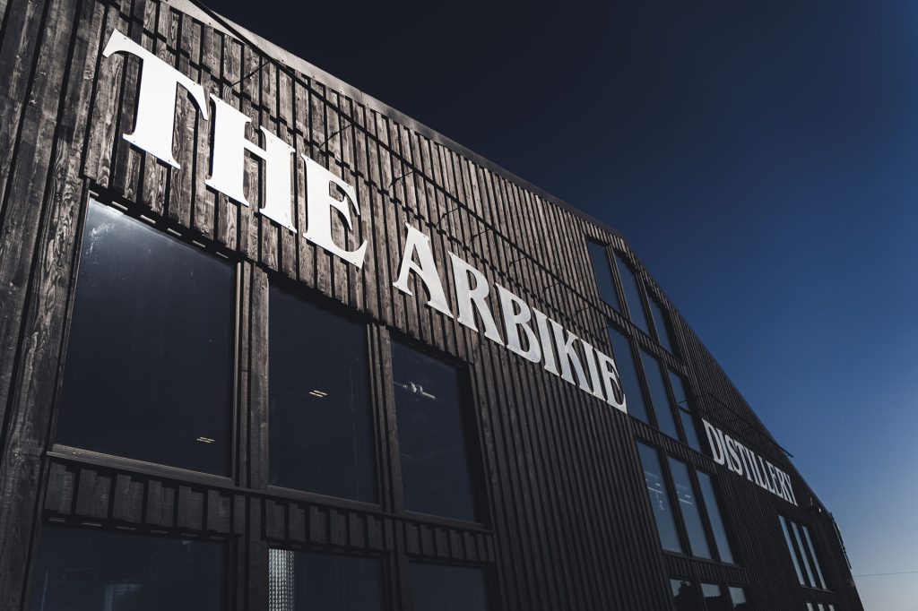 The image shows the exterior of a modern wooden building with large windows and the words The Arbikie Distillery in bold white letters against a clear blue sky.