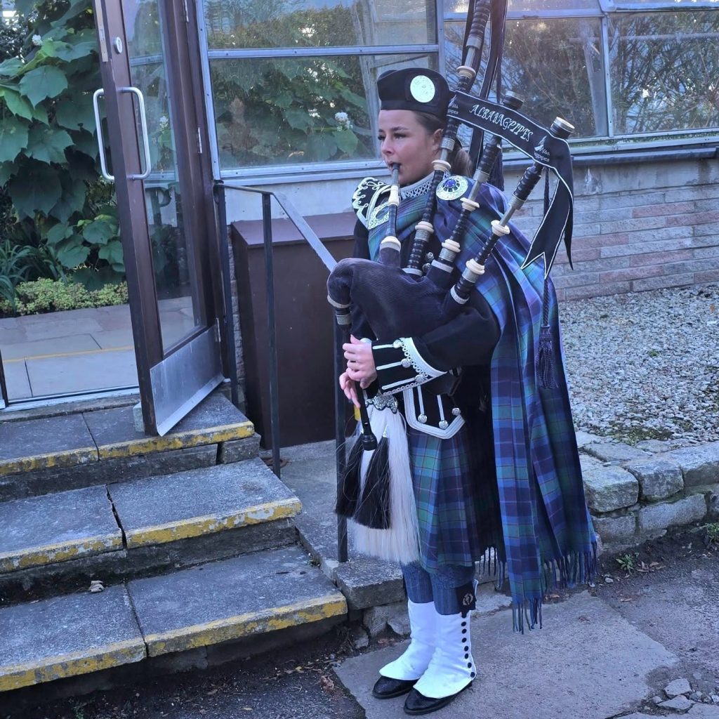 A woman dressed in traditional Scottish attire stands outside holding bagpipes. She wears a tartan sash, kilt, sporran, feather bonnet, and white shoes. The background features stone steps, greenery, and a glass door.
