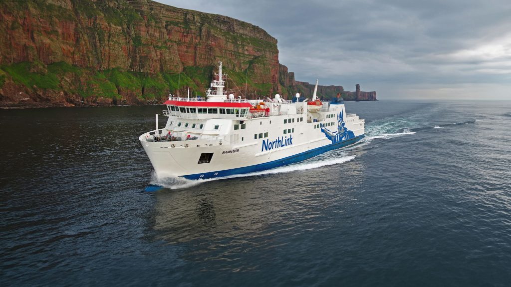 A NorthLink ferry sails on calm water near steep, green cliffs under a cloudy sky, leaving a trail in the sea behind it.