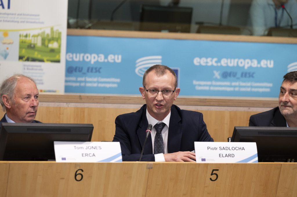 Three men in suits sit at a conference table with nameplates, microphones, and blue signs behind them reading europa.eu. The man in the middle, speaking, is identified as Piotr Sadlocha.