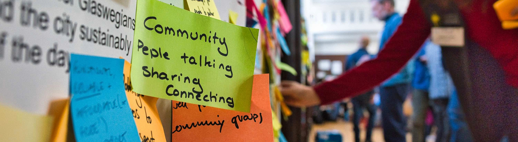 Colorful sticky notes on a wall with handwritten words like Community, People talking, Sharing, and Connecting. A person reaches toward the notes in a busy, indoor setting with other people in the background.