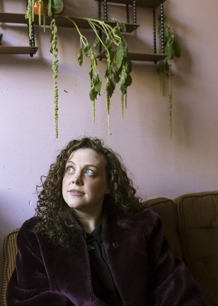 A woman with curly hair in a dark coat sits on a brown couch, looking up thoughtfully. Behind her, dried green plants hang upside down from shelves on a light purple wall.