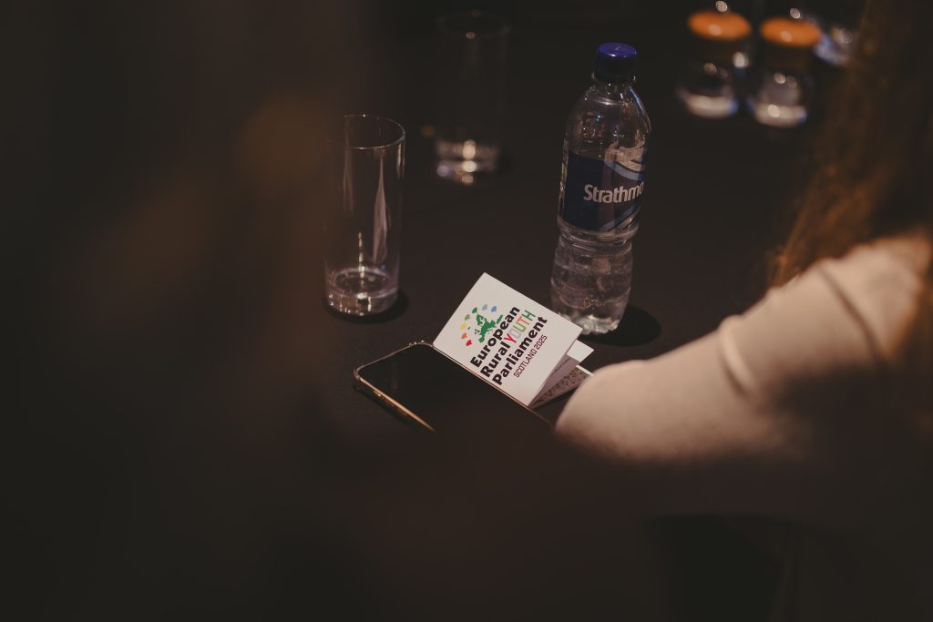 A person sits at a table with a water bottle, empty glasses, a phone, and a card labeled European Rural Parliament. The scene is dimly lit and focused on the card and bottle.