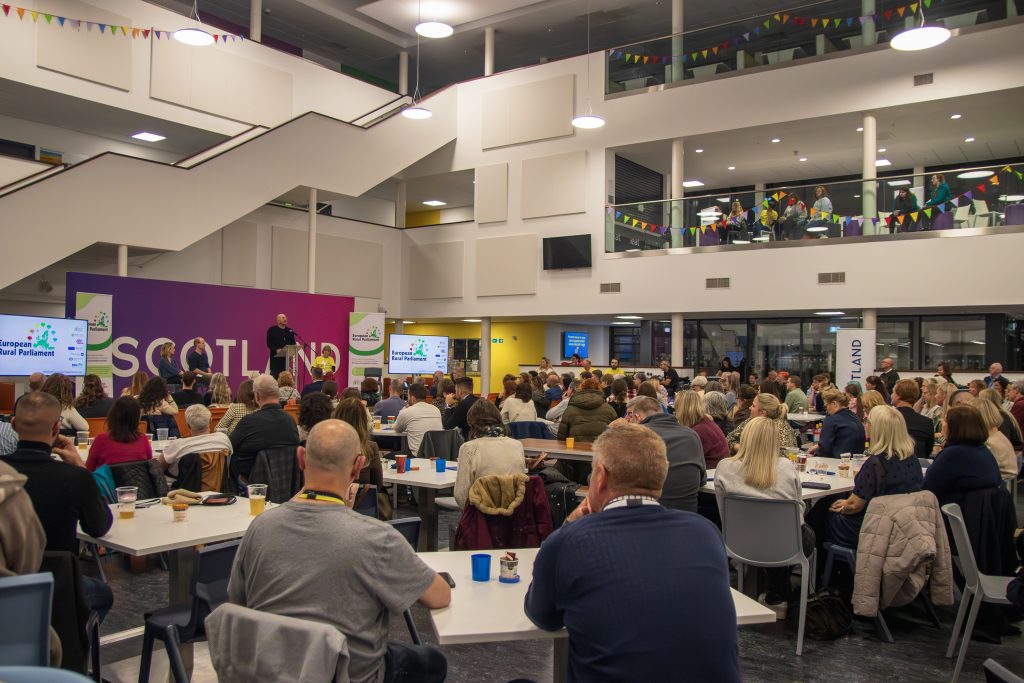 A large group of people sit at tables in a spacious, modern hall, listening to a speaker on stage in front of a purple backdrop that reads SCOTLAND. Two screens display presentation slides.