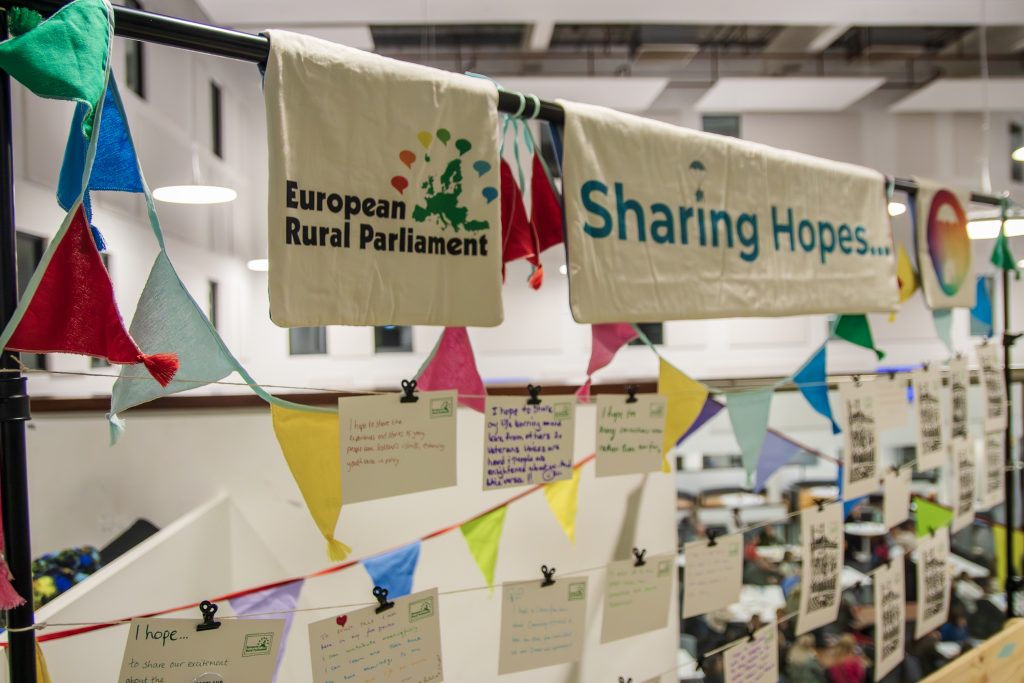 Colorful bunting and signs reading European Rural Parliament and Sharing Hopes... hang above handwritten notes clipped to a line in a bright indoor event space.