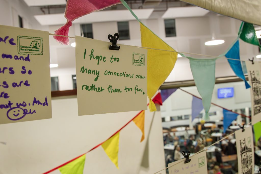 A handwritten note clipped to a string of colorful bunting reads, I hope too many connections occur rather than too few, displayed in a lively indoor setting with people in the background.
