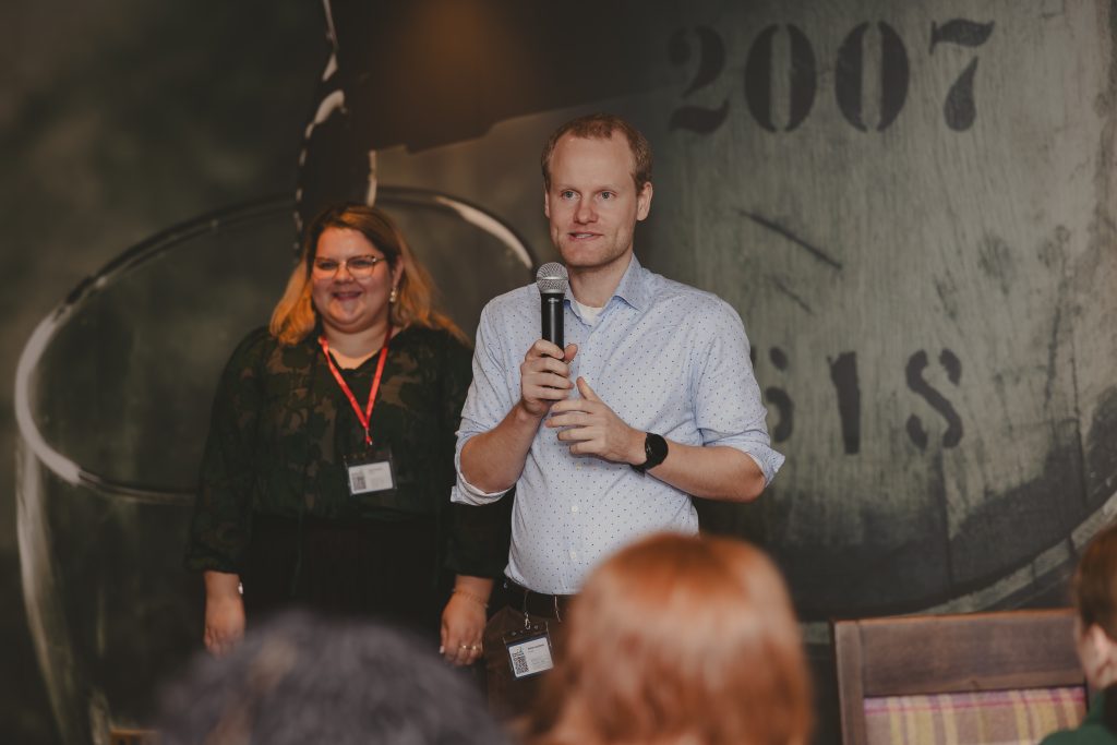 A man holding a microphone speaks to an audience, while a woman with a name badge stands smiling behind him. A dark wall with large numbers is in the background.
