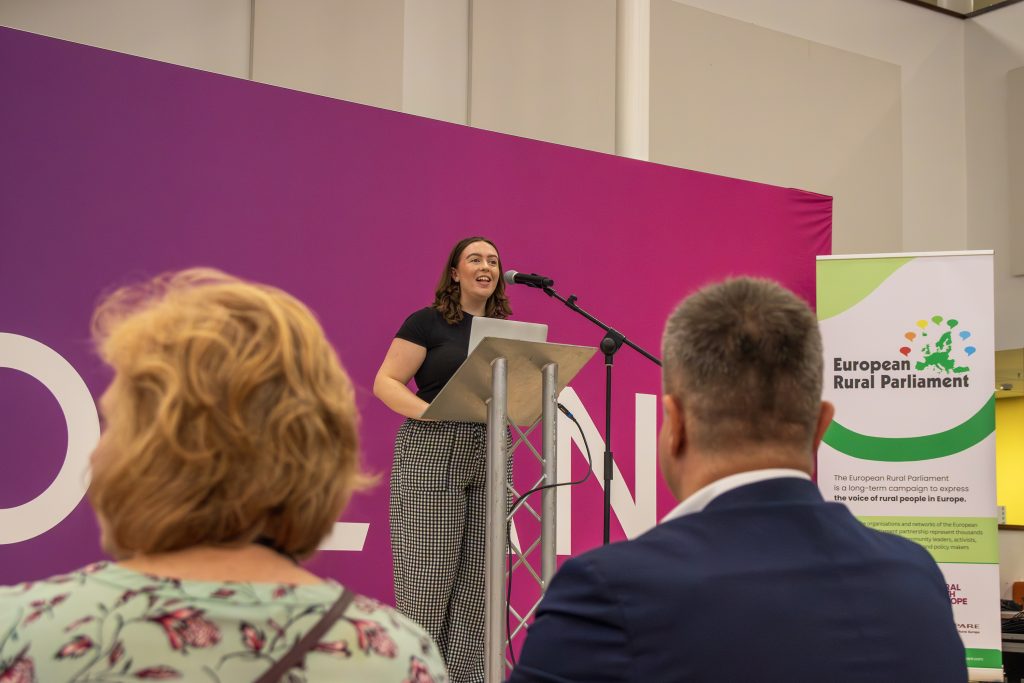 A woman stands at a podium speaking into a microphone at an event. People sit and listen in the foreground. A banner for the European Rural Parliament is visible on a purple and pink background.