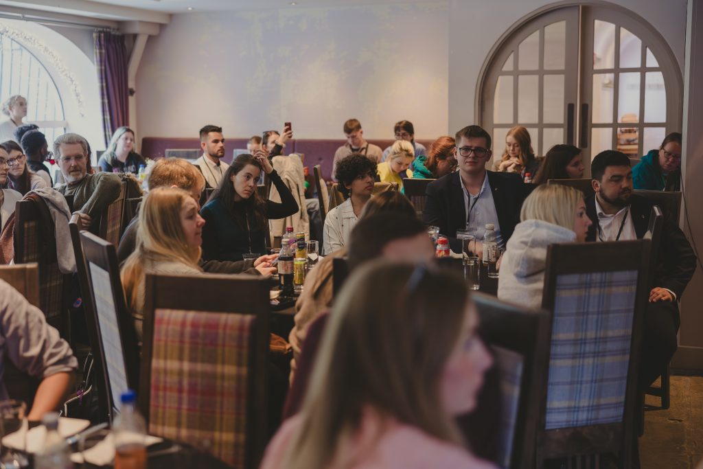 A group of people sit at tables in a well-lit room, attentively listening to something off-camera. Some are taking notes or using their phones, and drinks are on the tables. The atmosphere appears casual yet focused.