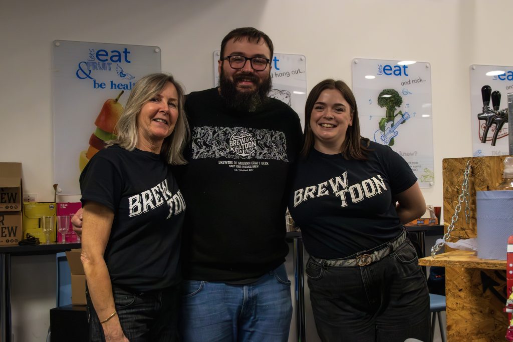 Three people stand together smiling indoors. The two women on either side wear matching Brew Toon t-shirts, while the man in the middle wears glasses and a black shirt. Posters and various objects are visible in the background.