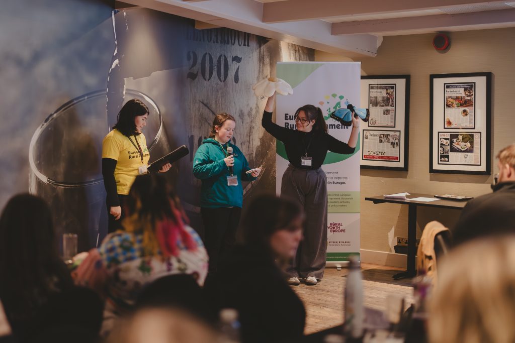 Three women stand at the front of a room presenting; one holds up her arms with gloves on. The audience sits facing them. A banner, framed photos, and a mural are visible in the background.