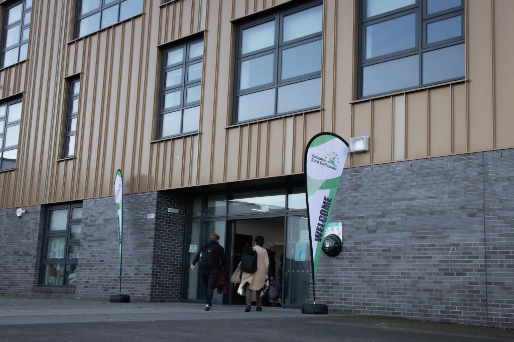 Two people walk into a modern building with large windows and brown paneling. Welcome flags and signs by the entrance suggest it is a special event or open day.