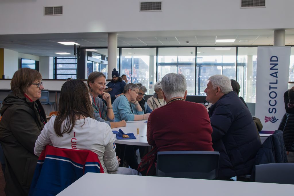 A group of people sit around a table in discussion in a modern, bright room with large windows. A Scotland banner is visible in the background.