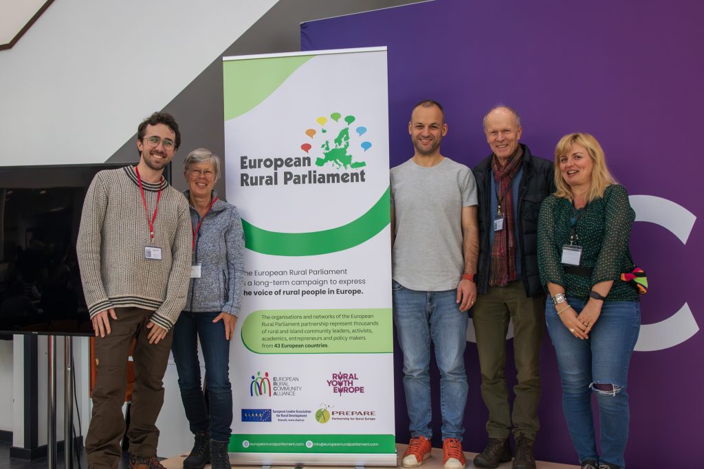 Five smiling people stand beside a European Rural Parliament banner in front of a purple wall; they wear conference badges and casual clothes, appearing at an event or meeting.