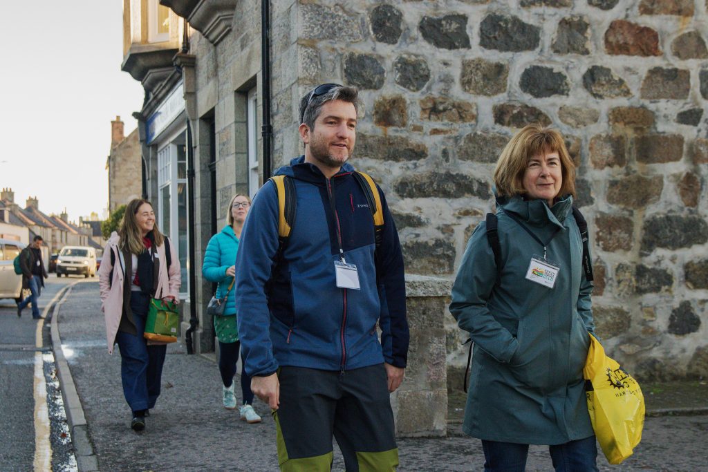 A group of people wearing jackets and name tags walk along a stone building on a sunny day; two people in front hold yellow bags, with more people and buildings visible in the background.