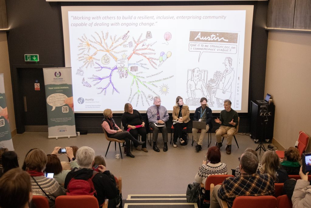 A panel of six people sit on stage in front of an audience, discussing community resilience. Behind them, a screen displays a colorful mind map and a cartoon. Audience members listen and take notes.