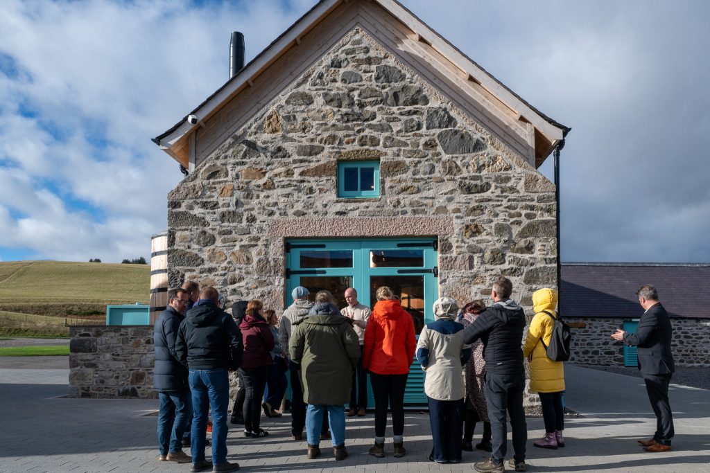 A group of people wearing jackets gather in front of a stone building with teal window frames on a sunny day, with grassy hills and blue sky in the background.