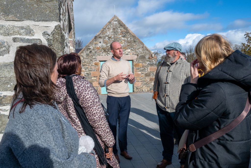 A man speaks to a small group of people outside near stone buildings on a sunny day, while the group listens attentively.