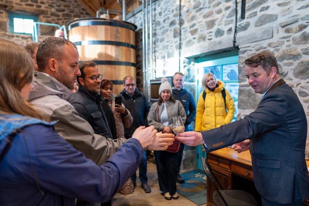 A group of people in winter clothing stand inside a stone-walled distillery as a man in a suit hands a glass of spirits to a guest for tasting; others watch and take photos.