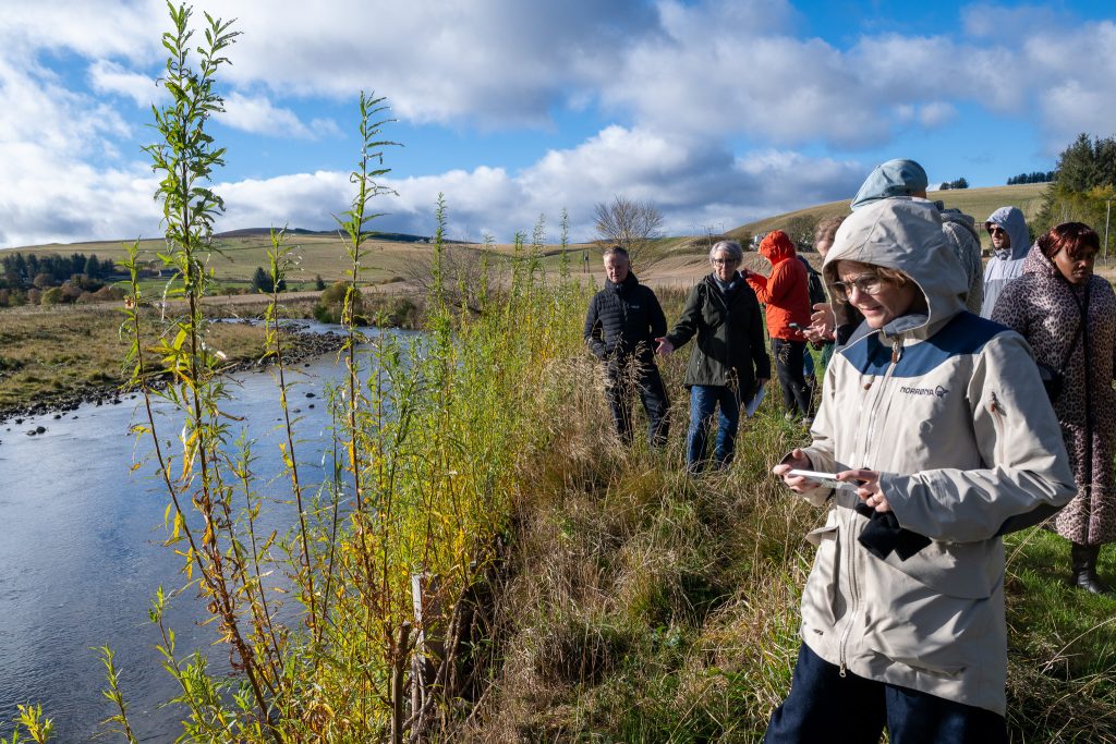 A group of people in jackets stands on a grassy riverbank under a partly cloudy sky, observing the water and taking notes. Rolling hills and trees are visible in the background.