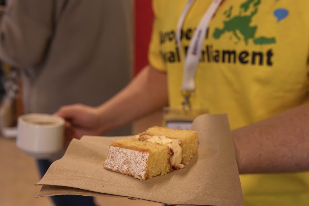 A person wearing a yellow European Parliament shirt holds a slice of cake on a napkin in one hand and a cup of coffee in the other. Another person is blurred in the background.