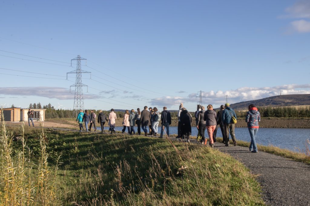A group of people walk along a path beside a body of water on a sunny day, with grassy fields, trees, and power lines in the background.