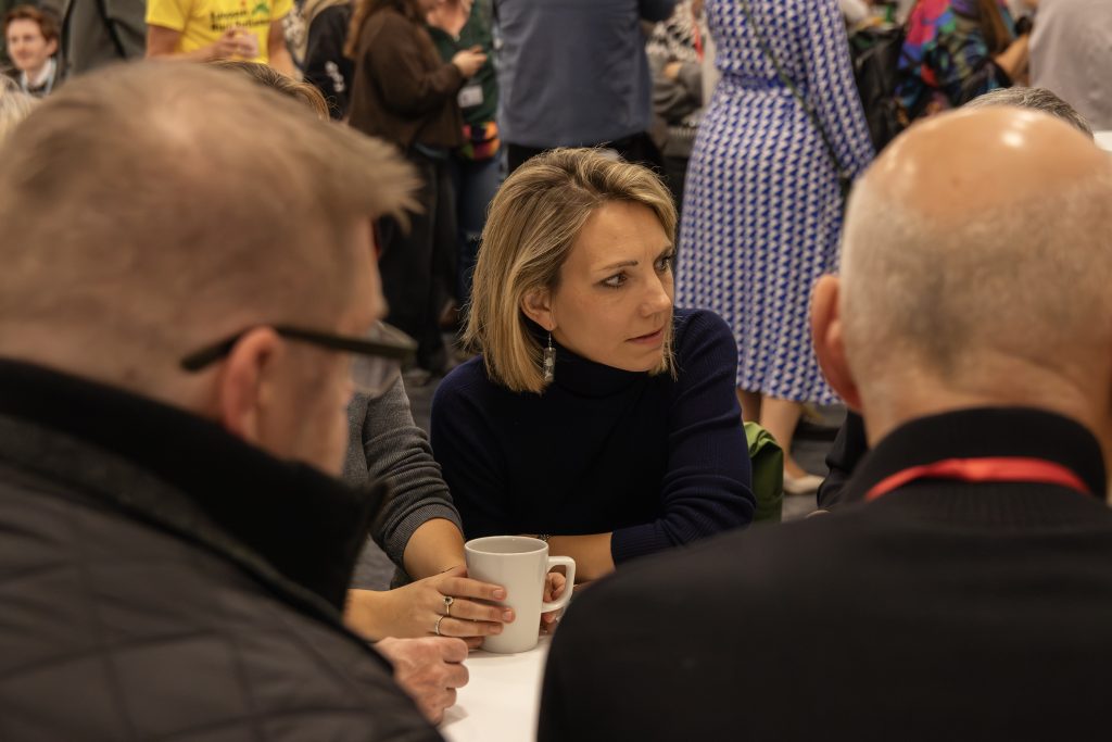 A woman holding a white mug sits at a table, engaged in conversation with three men in a busy indoor setting. This lively scene captures the spirit of connection and collaboration that defines our About Us story.