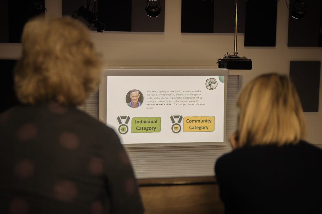 Two people sit facing a screen showing an award slide with a photo, text, and icons for “Individual Category” and “Community Category” medals. The scene is softly lit with an educational or presentation setting.