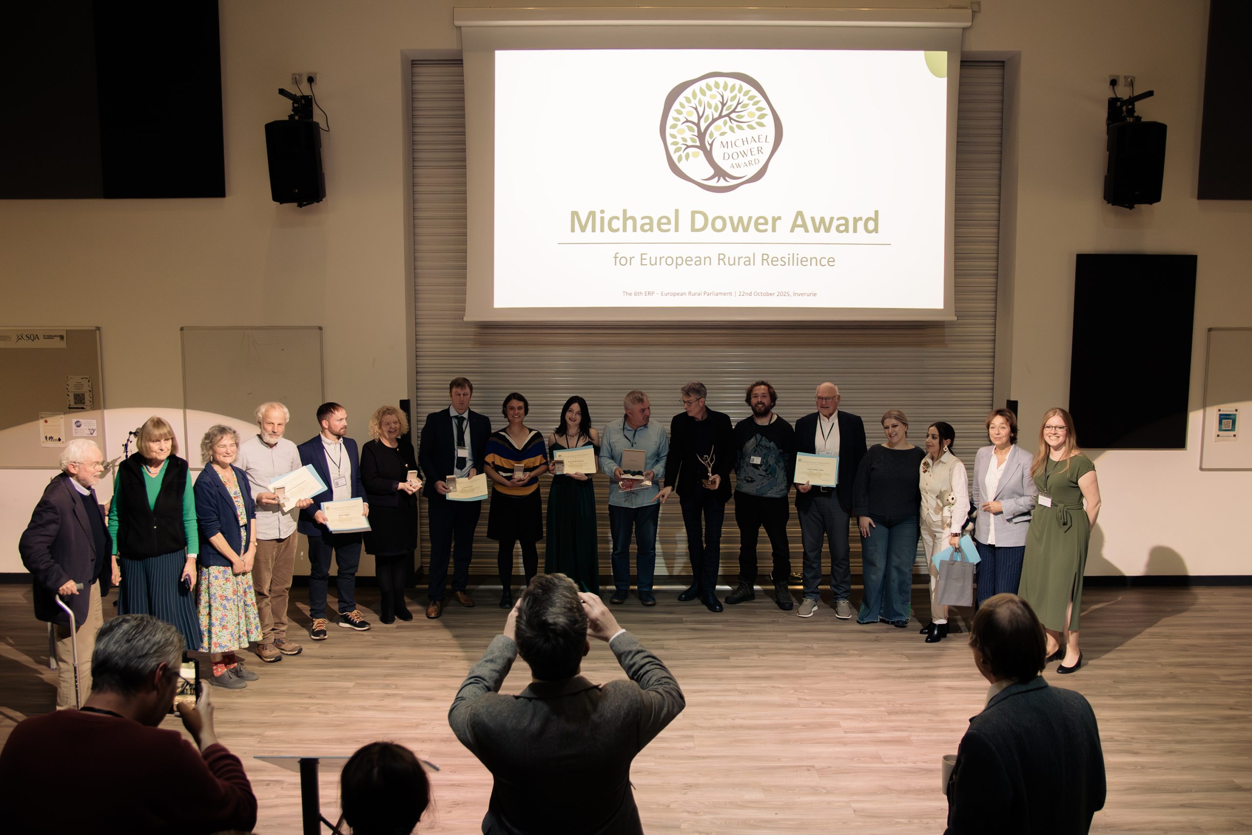 A group of people stands on a stage holding certificates during the Michael Dower Award for European Rural Resilience ceremony. An audience member takes photos as a presentation slide is displayed in the background.