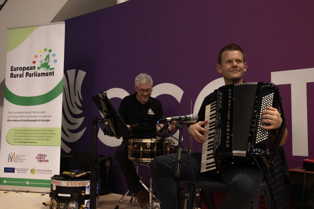 Two musicians perform indoors; one plays an accordion and smiles, while the other plays a snare drum. A banner for the European Rural Parliament stands in the background. Musical equipment is on the floor nearby.