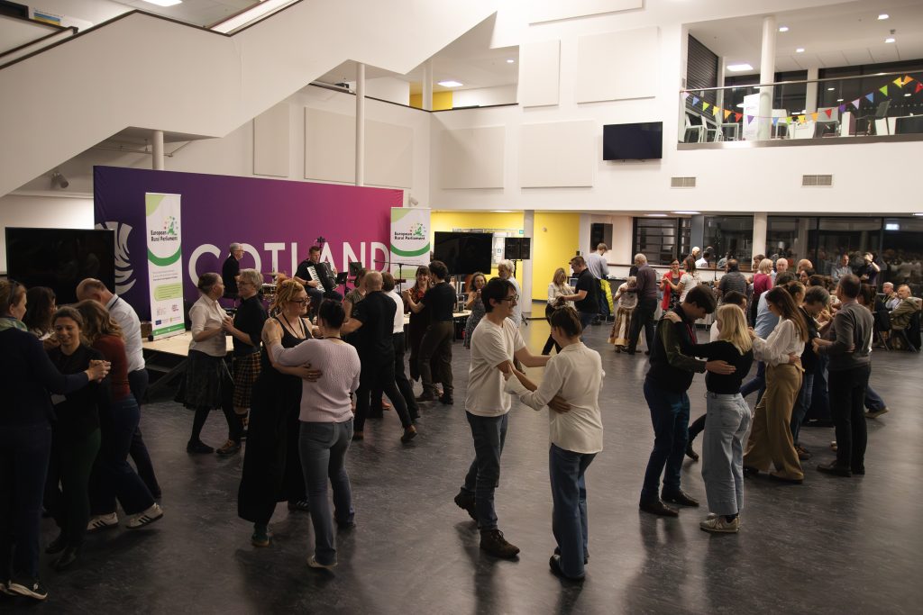 People dancing in pairs in a brightly lit indoor hall with modern decor. Colorful banners and a large GOTLAND sign are visible on the wall. Some people are seated, watching the dancers.