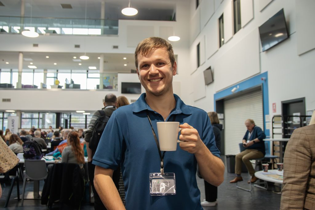 A smiling man in a blue polo shirt holds a white mug in a busy indoor setting with people seated at tables and talking in the background. He wears a lanyard and stands in a modern, well-lit building.