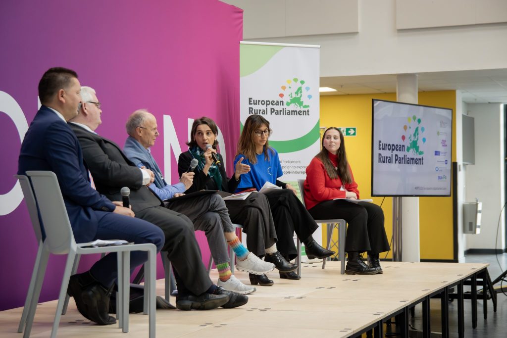 Six people sit on a stage in discussion at the European Rural Parliament event. One woman speaks into a microphone, while others listen. The backdrop is pink, and there is a screen and banner with the event logo.