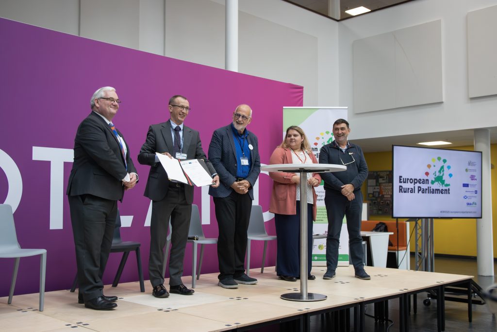 Five people stand on a stage at the European Rural Parliament event. One man is holding and presenting a certificate while others smile. A screen and event banner are visible in the background.