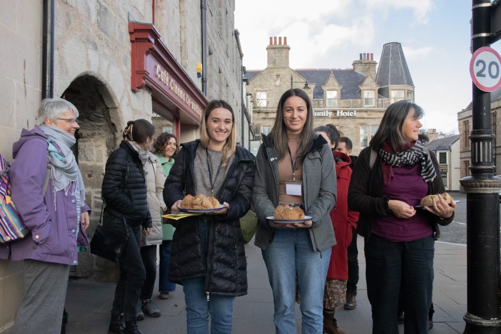 A group of people stand on a street in front of stone buildings. Two women in the front hold plates with bread, and others behind them are talking and smiling. It appears to be a chilly day, as everyone is wearing jackets.