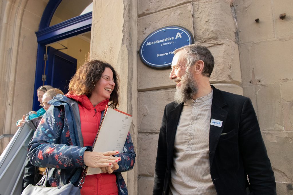 Two people stand outside a stone building, smiling and talking. The woman holds a folder and wears a red jacket, while the man has a beard and wears glasses. A blue Aberdeenshire Council sign is visible on the wall behind them.