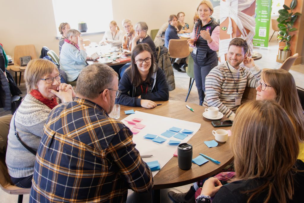 A group of people sit around a table covered with sticky notes, engaging in a lively discussion during a workshop. Several other groups are visible in the background, with coffee cups and papers on the tables.
