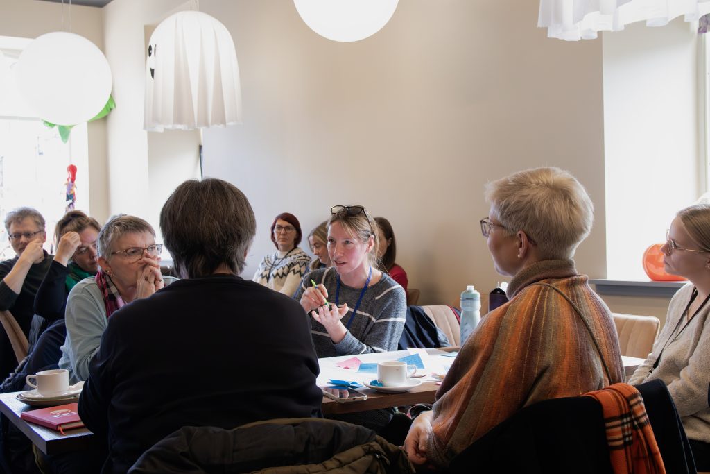 A group of people sit around a table in a well-lit room, engaged in conversation. Papers, notebooks, and cups are on the table, and some people are listening intently while one woman in the center is speaking and gesturing.