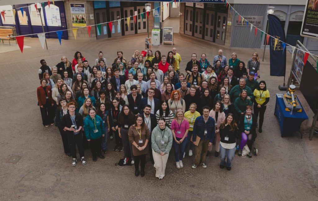 A large group of people pose together indoors under colorful bunting, smiling up at the camera. They are standing on a tiled floor in what appears to be an atrium or event space.