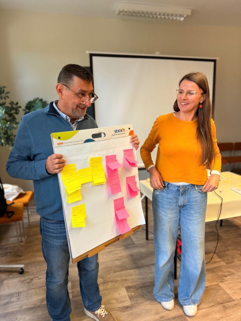 A man and woman smiling as they hold up a meeting pad with sticky notes organized into Strengths and Weaknesses columns.